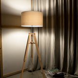 The Gaia Wood Floor Lamp - Light Brown, exuding rustic charm with its natural wood tripod, stands elegantly in a dimly lit room. Behind it, gray curtains drape over the window. A stack of magazines is neatly arranged on the floor to the right of the lamp.