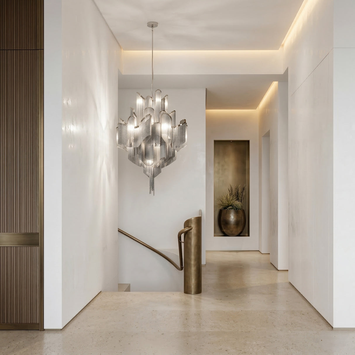 A modern, minimal hallway with cream walls and floor is highlighted by the Valora 19 Light Metal Chain Veil Chandelier in Nickel, a curved brass handrail on the staircase, a niche with a large vase of dried branches, and warm recessed lighting.