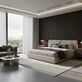 A modern bedroom features a large bed, neutral tones, floor-to-ceiling windows with a city view, sheer curtains, a gray sofa, and a round marble coffee table topped with decor and the Atria LED Alabaster Table Lamp – Brushed Brass.