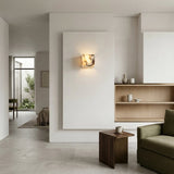 Minimalist living room with light beige walls, open wooden shelves, a small wooden side table with a mug, and a green sofa. Sunlight highlights the Alvara 1 Light Cube Veined Marble Wall Light in white & black near the open doorway.