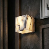 The Alvara 1 Light Cube Veined Marble Wall Light in White & Black glows on a dark textured wall, framed by wood trim and partially visible artwork.