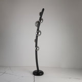 The Cordaro 6 Light Leather Floor Lamp - Black features a modern curved black rod with six metallic spheres, standing on a round black base placed on a cracked white surface.