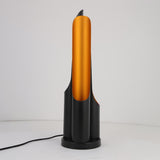 The Stealth Table Lamp - Brass & Black features a sleek cylindrical brass center and a matte black base, standing on a white surface with its black power cord trailing to the side.