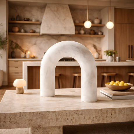 A modern kitchen with wooden cabinets and marble accents features the Arcella LED Sculpted Arch Alabaster Table Lamp – Soft White on a stone table, while a bowl of lemons and pendant lights create a warm, cozy atmosphere.