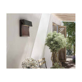 Sisal A/01 LED outdoor wall light in brown, woven fibre panel beneath an angled hood, mounted on an exterior wall near a pergola and plants.