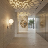 Modern hallway with light stone walls and floors, featuring the Pallo Wall Light - White with an opal glass diffuser, geometric wall decor casting patterned shadows, mirrors, and large windows revealing an outdoor tree for a bright, minimalistic feel.