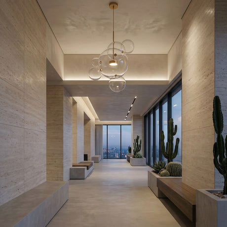 A modern hallway with beige stone walls and floors, large windows showing a city view at dusk, minimalist benches, cactus plants, and the Bubble LED Large Glass Pendant Light in gold hanging from the ceiling.