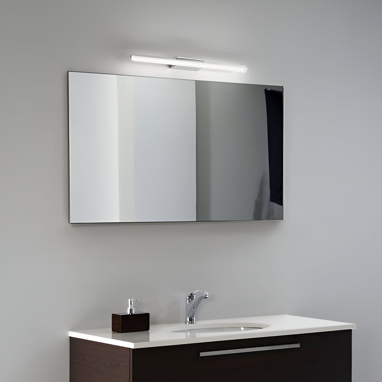 In a minimalist bathroom setting, a Luxix LED Bathroom Mirror Light 11W 3000K in chrome, measuring 42cm, casts a warm glow on the light gray wall. Below it sits a dark wood vanity with a white countertop, boasting a modern faucet and compact soap dispenser.