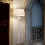 The Alloy Floor Lamp - White features a translucent, sculpted base and a large white shade that offers ambient lighting, perfect for adding a touch of modern elegance to an interior hallway accentuated by a dark wooden door and decorative stone arches with balustrades.