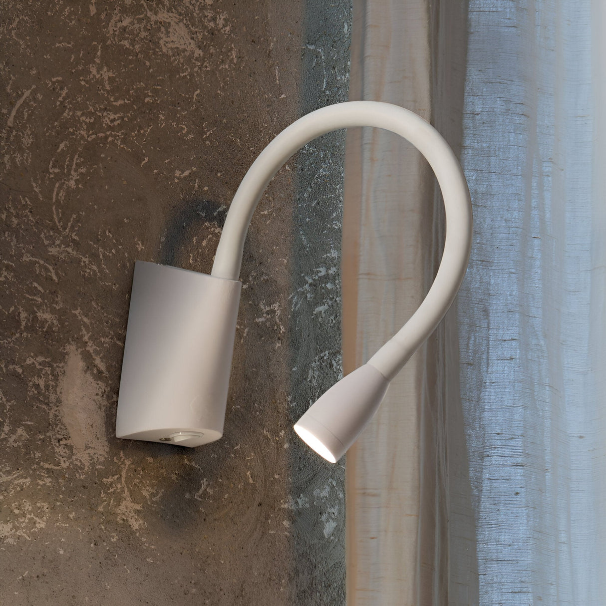 A contemporary Crown LED Wall Light in white, featuring a curved arm, is elegantly installed on a textured brown wall. Its LED bulb emits a focused beam of light, and the aluminium finish enhances its sophistication against a backdrop of sheer white curtains.