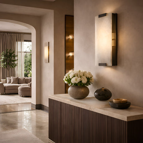 A modern hallway showcases a marble-topped console with a vase of white flowers and decorative bowls, softly lit by the Virelux LED Faceted Stone Alabaster Wall Light in Soft White & Black, opening into a cozy living room with neutral furniture and large windows.