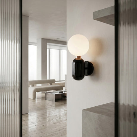 The Pallo Wall Light - Black with an opal glass diffuser is mounted on a white wall in a minimalist living room with light floors, neutral furniture, and large windows.