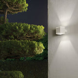 The Opus Square Outdoor Wall Light in white, featuring Pyrex glass, illuminates a textured wall at night and casts a soft glow. To the left, lush green trees with dense foliage stand out against the dark sky, creating a serene outdoor ambiance.