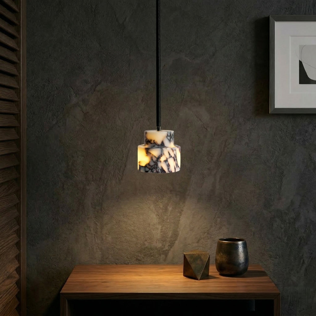 The Valmere 1 Light Veined Marble Single Pendant Light in white and black hangs above a wooden table with a metallic geometric object and ceramic cup, set before a dark textured wall decorated with framed art.