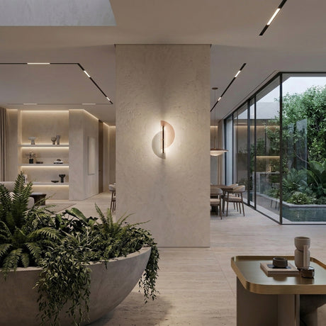 A modern, minimalist living space with light stone walls, built-in shelves, a large planter of green ferns, floor-to-ceiling garden windows, and the Papillon LED Wall Light - Silver & Bronze adding a sleek contemporary touch.