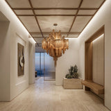 A modern, minimalist hallway features the Valora 19 Light Metal Chain Veil Chandelier in champagne, light stone flooring, built-in wood seating, a cactus planter, abstract wall art, and floor-to-ceiling windows overlooking the ocean.