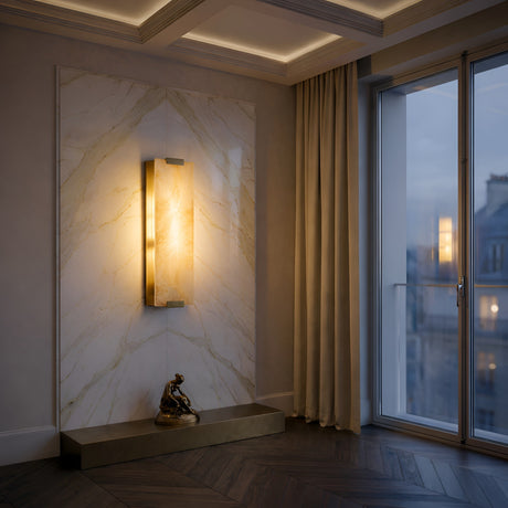 A modern interior with a marble wall panel showcases the Virelux LED Faceted Stone Alabaster Wall Light in soft white and brass, a gold sculpture on a pedestal, floor-to-ceiling curtains, and a large window overlooking city buildings at dusk.