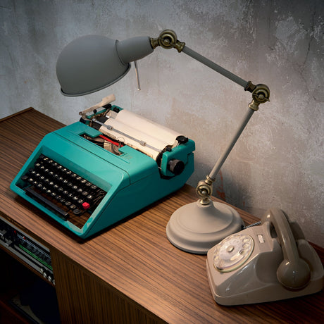 On a wooden desk, a vintage teal typewriter is placed beside a classic gray rotary dial telephone and the Neo Table Lamp in gray. The textured, rustic wall in the background creates a charming contrast between these timeless items and the subtle modernity of the workspace lighting.