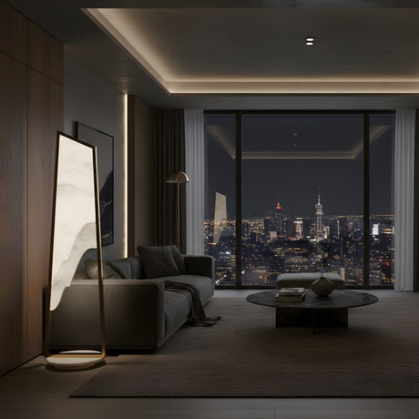 A modern living room at night showcases a gray sofa, round coffee table, and city views, accented by floor-to-ceiling windows and the Atria LED Small Alabaster Floor Lamp in Brushed Brass beside a Spanish alabaster panel.