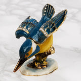 Perched Kingfisher Metal Trinket Box with hinged lid, glossy blue finish, yellow breast, gold feather detailing and crystal-studded wings, on a marbled white background.