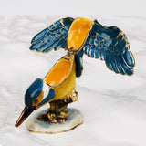 Perched Kingfisher Metal Trinket Box with hinged wings open, revealing a yellow-orange interior, blue and gold enamel feathers on a white marbled surface.
