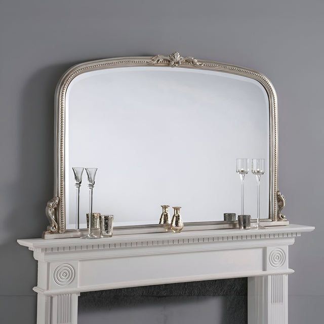 The Mist Overmantle Wall Mirror Gold - 122x79cm, featuring an ornate frame with a gold finish, is positioned above a white mantel. The luxurious classic arrangement includes three glass bottles, two champagne flutes, and small candles, all elegantly displayed against a gray wall.