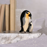 Penguin & Chick handpainted metal trinket box, black and white with yellow chest wash and sparkling silver-tone wings, shown on a marble surface beside books.