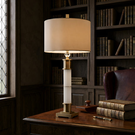 The Alverin 1 Light Segmented Alabaster Table Lamp in Brass & Soft White rests on a wooden desk in a classic library with old books, a leather chair, and wood shelves. Sunlight pours through the leaded glass window.