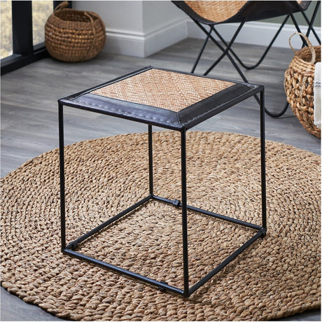 Ezio 41cm stool with black leather-trimmed rattan seat and black iron cube frame, shown on a jute rug in a room with woven baskets and a chair.