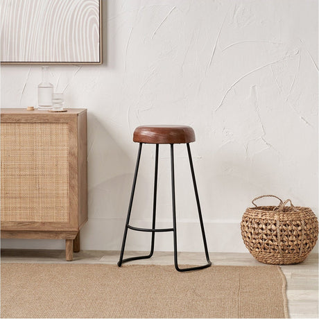 Cosimo Vintage Brown Leather & Black Iron Bar Stool, 77cm, with round leather seat and slim splayed metal legs, shown in a neutral room beside a wooden sideboard and woven basket.