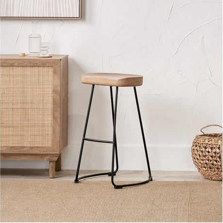 Laselle Ash Wood & Black Metal Bar Stool, 70cm, with a shaped wooden seat on angled black metal legs and footrest, shown beside a rattan sideboard in a dining setting.