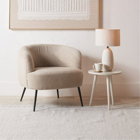 Aviana armchair in taupe teddy fabric with curved back and black metal legs, styled beside a small side table and lamp in a light living room setting.