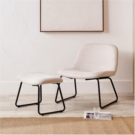 Chiara lounging chair and matching footstool in natural bouclé with black metal sled legs, shown in a light-toned living room beside a folded magazine and glass.