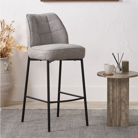 Enya bar stool with light grey chenille quilted back and seat on slim black metal legs with footrest, shown in a room beside a small round side table.