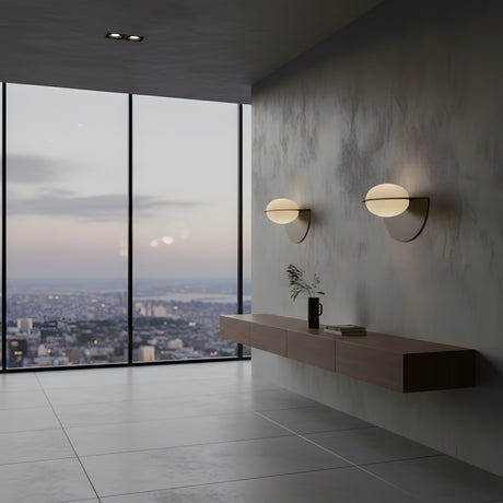 A modern minimalist interior featuring a floating wooden shelf, a vase, the Althia LED Alabaster Wall Light in matte black, and floor-to-ceiling windows with a cityscape view at dusk.