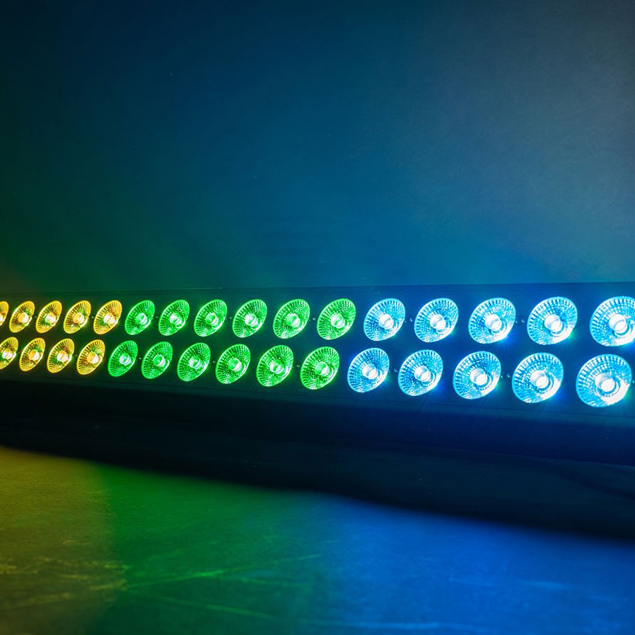 QTX DuplexBar AVSL LED wall bar with two rows of 24 RGBA lenses lit in amber, green and blue against a hazy blue backdrop.