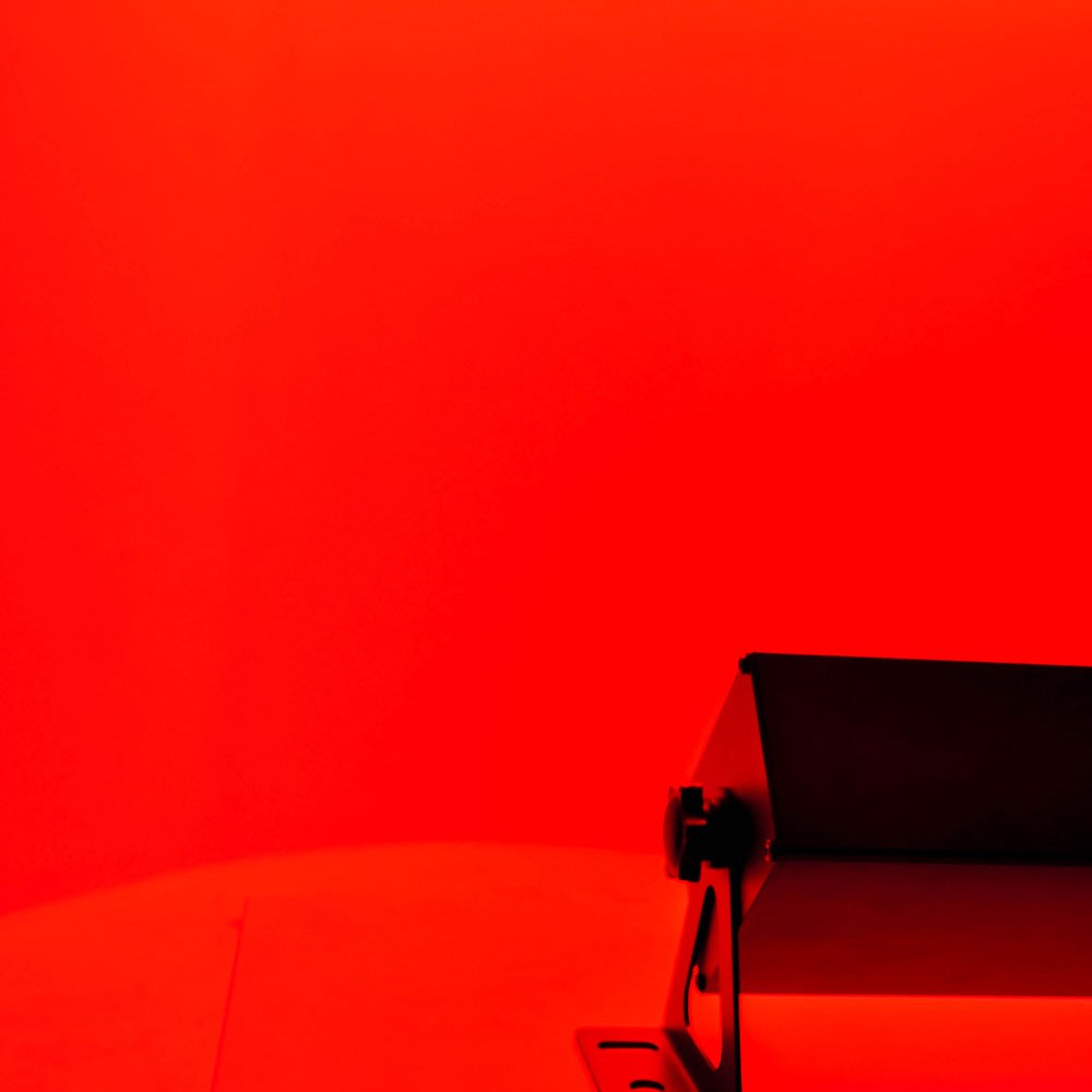 QTX DuplexBar LED wall bar by AVSL with tilt bracket, shown end-on under intense red wash lighting against a matching red backdrop.