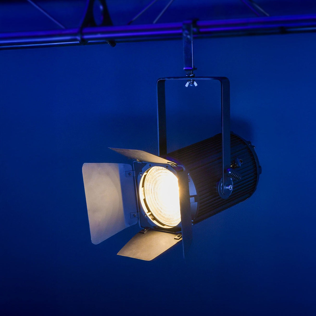 Citronic MFZ100-W mini Fresnel zoom LED stage light illuminated warm white with barn doors, mounted on a truss against a blue-lit backdrop.
