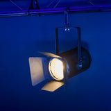 Citronic MFZ100-W mini Fresnel zoom LED stage light illuminated warm white with barn doors, mounted on a truss against a blue-lit backdrop.