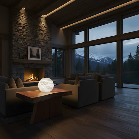 A cozy living room with a stone fireplace, two sofas, a Noreva 1 Light Sculpted Globe Alabaster Table Lamp – Soft White on a wooden table, and large windows revealing snowy mountains and pine trees at dusk.