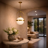 The Calis Alabaster Single Pendant Light in Brushed Brass & Soft White hangs above a round marble table with white flowers in a softly lit hallway, with a cozy living room and large windows seen in the background.