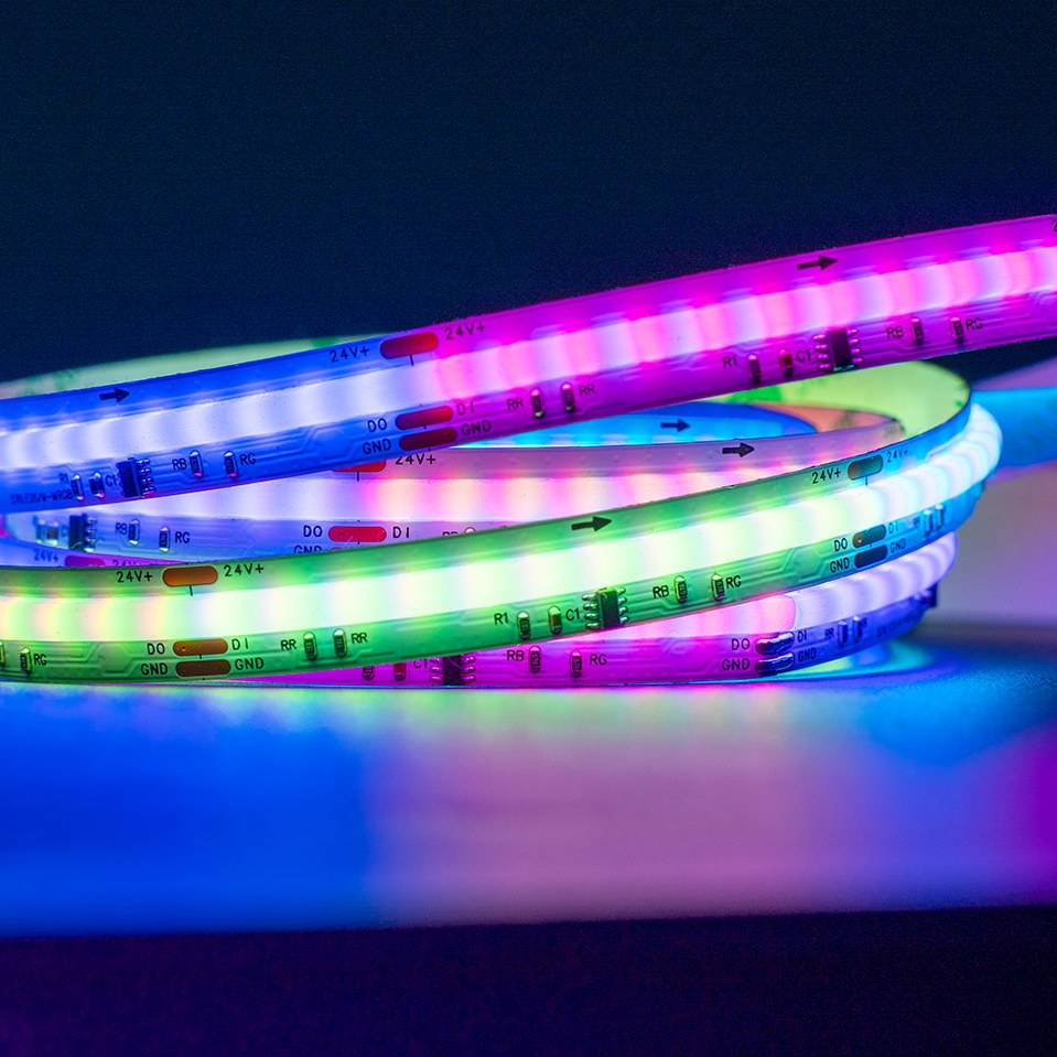 Coiled RGB LED tape lit in pink, green, blue and white along a flexible strip marked with 24V, DO, DI and GND solder pads on a dark background.