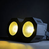 AVSL QTX 2x50W LED wash light with two round white COB lenses in a black metal housing, side mounting bracket, illuminated warm against a dark background.