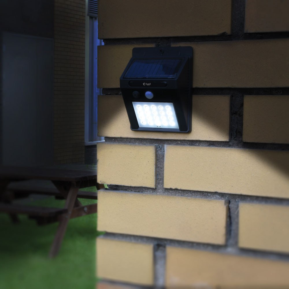 LYYT solar LED wall light in black with illuminated LED panel and motion sensor, IP44 rated, mounted on a yellow brick exterior wall at dusk.