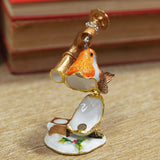 Hinged metal trinket box shaped as a robin with orange breast drinking from a gold tap, set on a white snowy base with holly, on a wooden surface.