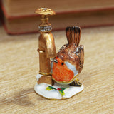 Robin Drinking from Tap hinged metal trinket box, featuring a brown-winged robin with orange breast beside a gold tap on a white snowy base with holly, shown on a wooden surface.