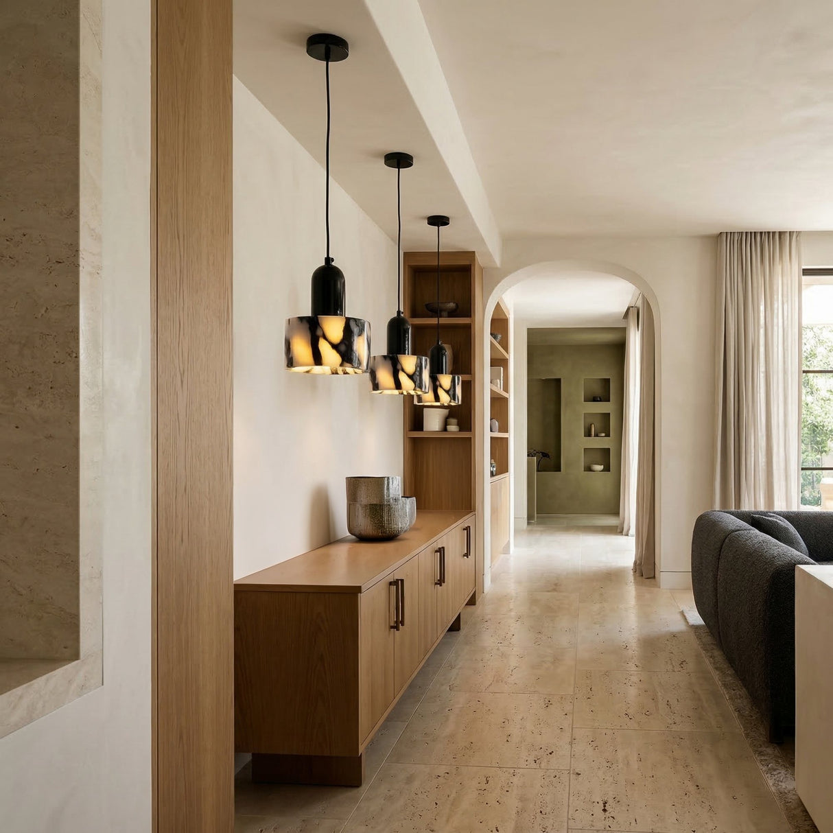 A modern hallway with beige stone floors, wooden cabinets, built-in shelves, and Valere 1 Light Veined Marble Single Pendant Light - White & Black above the cabinets. The space leads to an archway with large windows and light curtains beyond.