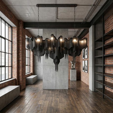A modern loft features exposed brick, large windows, wooden floors, and a striking Valora 15 Light Metal Chain Veil Suspension Light in black. Concrete accents and minimalist art enhance the industrial-style décor.