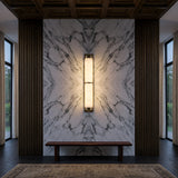 A symmetrical modern interior features a marble accent wall, vertical wooden panels, and a central Arelis LED Linear Alabaster Wall Light in Soft White & Brushed Brass. A simple bench sits below large windows, with part of a rug visible on the floor.