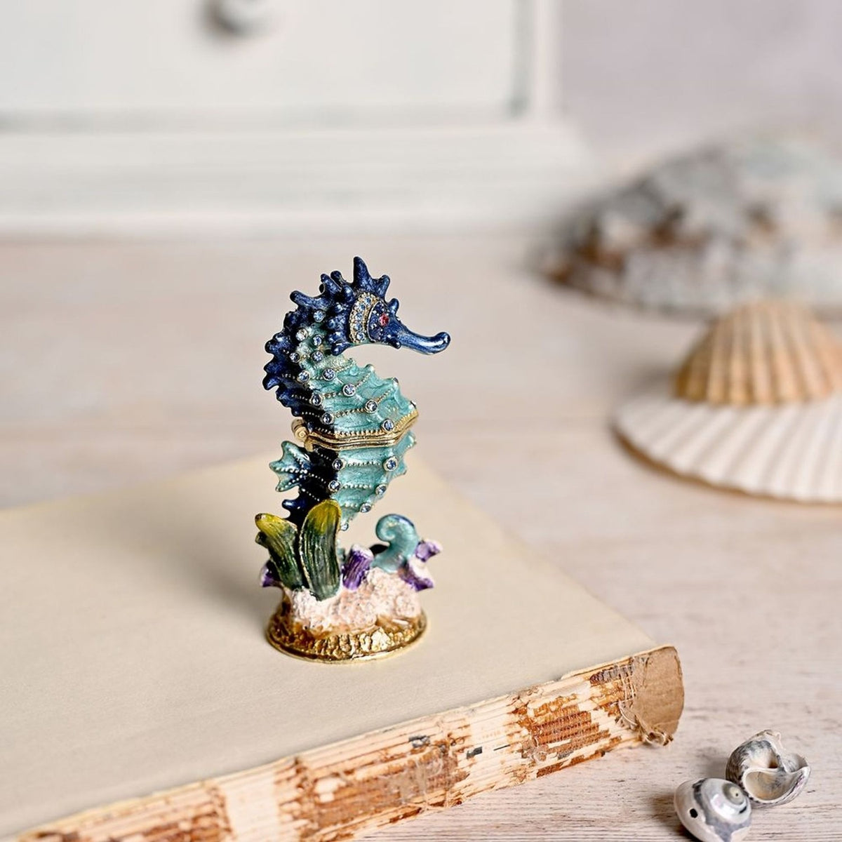 Sea Horse hand painted metal trinket box with hinged lid in turquoise and navy blue, gold base with seaweed and coral details, displayed on a book with shells in a softly blurred setting.